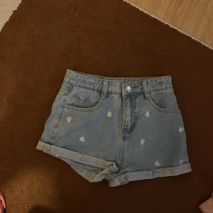 Blå jeansshorts med broderade blommor - Snygga blå jeansshorts från Shein med broderade vita blommor framtill. Modellen är högmidjad med klassiska fickor både fram och bak samt uppvikta benslut. Perfekt för dig som vill ha en chill och trendig sommarstil.