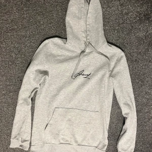 Ljusgrå hoodie med svart tryck - Säljer en ljusgrå hoodie från Drey med svart signaturtryck på bröstet. Tröjan har huva med snörning och en stor magficka. Perfekt för dig som gillar en enkel och stilren look.