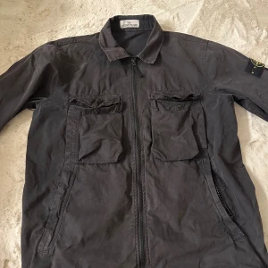 Svart overshirt från Stone Island - Svart overshirt från Stone Island med klassisk krage, två stora bröstfickor med lock och dragkedja framtill. Ikonisk patch på vänster ärm. Jackan är tillverkad i bomull och har en clean, urban look som passar streetwear-stilen. Inte använd den på ett tag som man kanske ser. Fraktar alltid samma dag som köp.