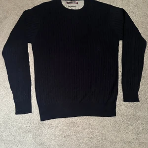 Mörkblå ribbad stickad tröja - Mörkblå stickad tröja från NP med ribbad struktur och rund hals. Tröjan har långärmade smala ärmar och en klassisk passform som funkar till jeans eller chinos. Perfekt för dig som gillar stilrena och enkla plagg.