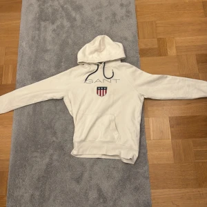 Vit hoodie från GANT med logga - Vit hoodie från GANT i storlek S med broderad logga och emblem på bröstet. Tröjan har huva med svarta snören, långärmad design och en klassisk känguruficka framtill. Mjuk och skön bomullskvalitet.