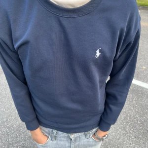 Mörkblå sweatshirt från Polo Ralph Lauren - Klassisk mörkblå sweatshirt från Polo Ralph Lauren med rund halsringning och broderad vit logga på bröstet. Tröjan har ribbade muddar vid ärmslut och nederkant. Perfekt för en clean och stilren look. Tröjan är använd en gång bara. Nypris 899kr, storlek S. Priset kan diskuteras vid snabb affär.