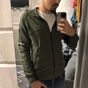 Olivgrön Nike track zip upp - Nike track jacket i olivgrönt med hel dragkedja, ståkrage och svarta detaljer. Jackan har två fickor med dragkedja och vita reflexdetaljer på ärmarna. Perfekt för träning eller chill, snygg och sportig look.