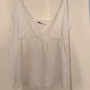 Vit spetsdetaljerad linnetopp Nelly - Supersöt vit linnetopp från Nelly med spetsdetaljer längs v-ringningen och nederkanten. Toppen har smala axelband, liten rosett framtill och är tillverkad i mjuk bomull. Perfekt för en romantisk look eller som layering-piece. Aldrig använd🥰