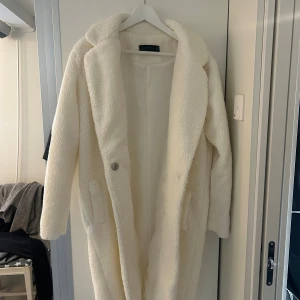 Vit teddyjacka från Dilvin Woman - Säljer en lång, fluffig teddyjacka i vitt från Dilvin Woman. Jackan har bred krage, stora fickor och stängs med knapp framtill. Perfekt för dig som vill ha en mysig och trendig look under kyliga dagar.