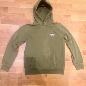 Olivgrön hoodie GANT - Olivgrön hoodie från GANT med broderad logga och sköld på bröstet. Klassisk känguruficka framtill och huva. Mjuk och skön bomullsblandning, perfekt för chill dagar. Snygg och enkel design som funkar till det mesta. Storlek 170 men sitter som s
