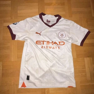 Manchester City Haaland bortatröja Puma - Snyggaste Manchester City bortatröja med Haaland 9 på ryggen. Vit med vinröda detaljer på krage och ärmslut, guld glänsande Puma-logga och Etihad Airways-tryck. Passar perfekt att träna i eller göra en snygg outfit i.