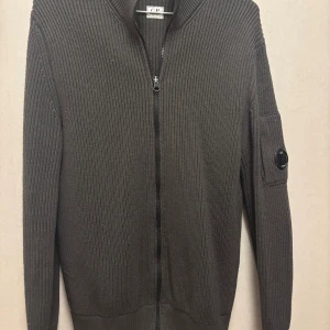 Grå stickad kofta från C.P. Company - Riktigt snygg grå stickad kofta från C.P. Company med dragkedja framtill och deras ikoniska linsdetalj på ärmen. Koftan är ribbstickad och har lång ärm, perfekt för lager på lager under kyliga dagar.                                                                                          Storlek 52 men sitter lite tajt runt midjan så passar M också. Köpt för 3500kr på NK 