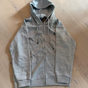 Grå hoodie från Polo Ralph Lauren - Snygg grå hoodie från Polo Ralph Lauren med dragkedja och huva. Klassisk broderad logga på bröstet och ribbade muddar. Perfekt för en chill och stilren look. Gjord i mjuk bomullsmix och har snörning i huvan.
