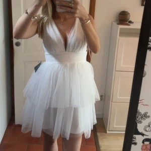 Vit  studentklänning  - Drömmig vit klänning med djup v-ringning både fram och bak. Volang, Klänningen har flera lager tyll som ger en fluffig och romantisk känsla. Midjan är markerad med ett brett band och klänningen stängs med dragkedja i ryggen. Perfekt studentklänning, aldrig använd 