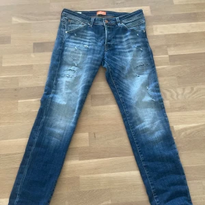 Blå slitna jeans från Jack & Jones - Säljer ett par blå jeans från Jack & Jones med snygga slitningar och ljusa tvättade partier. Jeansen har klassisk femficksdesign, raka ben och slim passform. Perfekta för dig som gillar en avslappnad och trendig look.
