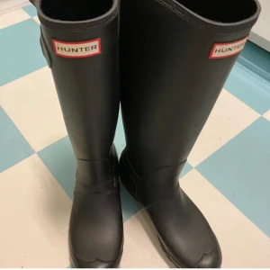 Svarta Hunter stövlar - Klassiska svarta stövlar från Hunter med rund tå och platt sula. Tillverkade i slitstark syntet, perfekt för regniga dagar. Stövlarna har Hunter-loggan framtill.