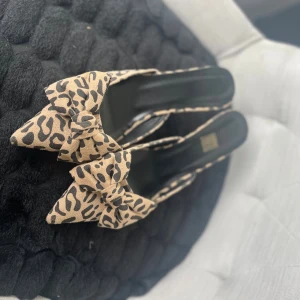 Leopardmönstrade pumps med rosett - Snygga pumps med spetsig tå i beige och svart leopardmönster. Skorna har en stor rosett framtill och platt sula med gulddetalj på hälen. Materialet är mocka och de har en elegant, feminin look som sticker ut.