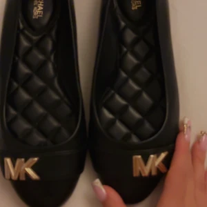 Svarta ballerinaskor Michael Kors - Snygga svarta ballerinaskor från Michael Kors med quiltad innersula och rund tå. Skorna har en platt sula och är dekorerade med guldfärgade MK-detaljer framtill. Tillverkade i skinn för en lyxig känsla och stilren look.