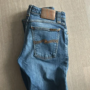 Blå jeans från Nudie Jeans - Snygga blå jeans från Nudie Jeans med klassisk femficksdesign och kontrastsömmar. Jeansen har en rak passform och är tillverkade i mjukt denimtyg av bomull. Bakfickorna har Nudie Jeans signatursöm och läderpatch i midjan. Jag är ungefär 180. 27/34 storlek