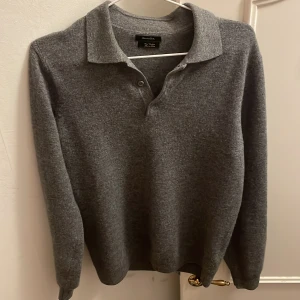 Grå kashmirpiké från Massimo Dutti - Mjuk och lyxig grå pikétröja i kashmir från Massimo Dutti. Klassisk krage, knäppning med två knappar och lång ärm. Perfekt för dig som vill ha en stilren och bekväm tröja med premiumkänsla.