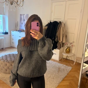 Grå offshoulder stickad tröja - Mysig grå stickad tröja med bred ribbad offshoulder-krage och långa ärmar. Tröjan har en loose fit och är perfekt för kyliga dagar. Den grova stickningen ger en trendig och avslappnad vibe.