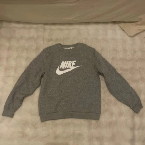 Grå Nike sweatshirt XL barn storlek  - Grå sweatshirt från Nike i storlek XL i barnstorlek, med klassisk vit Nike-logga framtill. Tröjan har rund halsringning och långa ärmar. Mjuk insida och ribbade muddar vid ärmslut och nederkant. Perfekt för chill dagar eller till träning.