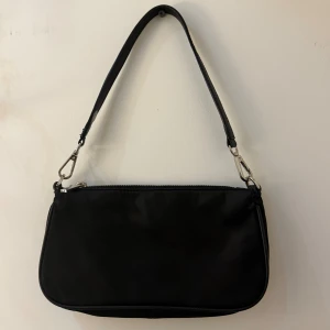 Svart axelväska från Zara - Stilren svart axelväska från Zara med avtagbar rem och silvriga metalldetaljer. Väskan har en klassisk rektangulär form och stängs med dragkedja. Tillverkad i syntetmaterial.