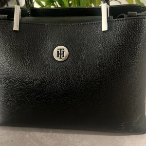 Svart handväska från Tommy Hilfiger - Stilren svart handväska från Tommy Hilfiger i präglat skinn med silverfärgade metalldetaljer och TH-logga framtill. Väskan har dubbla handtag och praktiska fack, perfekt för dig som vill ha en klassisk och snygg accessoar till vardagslooken.