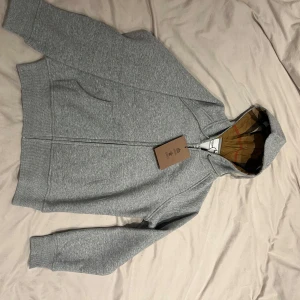 Grå Burberry hoodie med klassisk huva - Säljer en grå hoodie från Burberry med dragkedja och klassisk rutmönstrad insida på huvan. Tröjan har fickor framtill och är tillverkad i mjuk bomull med lite elastan för extra komfort. Perfekt för dig som gillar stilrena och exklusiva plagg. Den är storlek L men väldigt liten i storlek så mer s/m. Skriv vid fler frågor🥰