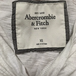 Vit twistad topp Abercrombie & Fitch XS - Supersöt vit topp från Abercrombie & Fitch i storlek XS. Toppen har en twistad detalj framtill, v-ringning och korta ärmar. Tillverkad i mjuk bomull, perfekt för en chill och stilren look.