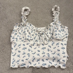 Vit blommig croppad linnetopp - Supersöt vit croppad linnetopp med blått blommönster och volangiga axelband. Toppen har snörning framtill och vågig kant nertill. Perfekt för en somrig look och lätt att matcha med jeans eller kjol.