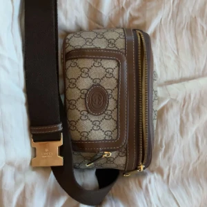 Gucci magväska med GG-mönster - Snygg magväska från Gucci med klassiskt GG-mönster i beige och brunt. Väskan har bruna skinn-detaljer, guldfärgad spänne och dragkedja. Justerbar axelrem i mörkbrunt tyg och praktiska fack framtill. Perfekt för dig som vill ha en ikonisk designer-accessoar.NY PRIS 11000kr
