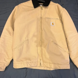 Beige Carhartt bomberjacka M - Säljer en beige Carhartt bomberjacka med svart krage i manchester. Jackan har dragkedja framtill, två snedställda fickor och en bröstficka med Carhartt-logga. Insidan är quiltad för extra värme och passformen är normal. Perfekt för höst och vinter.