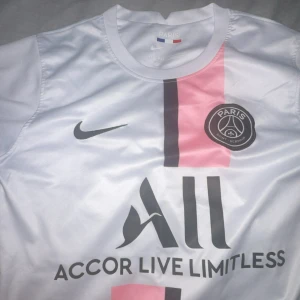 PSG vit och rosa fotbollströja Nike - Paris Saint-Germain fotbollströja i vitt med rosa och svart vertikal rand, officiellt klubbmärke och Nike-logga. Tillverkad i lätt och ventilerande Dri-FIT-material. Snygg design med trycket 'Accor Live Limitless' på bröstet.