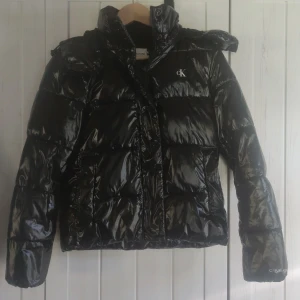 Svart pufferjacka Calvin Klein Jeans XS - Svart glansig pufferjacka från Calvin Klein Jeans med huva och dragkedja. Jackan har quiltad design, CK-logga på bröstet och Calvin Klein Jeans-tryck på insidan. Perfekt för kalla dagar och har en riktigt snygg streetstil.
