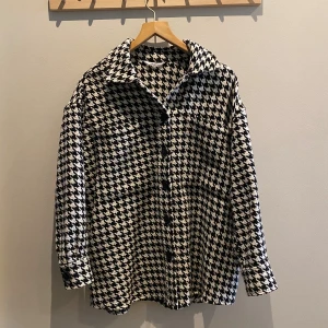 Jagcka från Ellos i svartvitt - Snygg jacka/overshirt från Ellos med klassiskt svartvitt mönster. Modellen är loose fit med stora krage, knappar framtill och långa ärmar med manschett. Storleken är 38-40. 