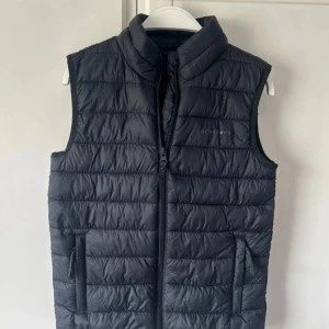 Svart dunväst från Ocean Works - Snygg svart dunväst från Ocean Works med quiltad design och hög krage. Västen har dragkedja framtill och två fickor med dragkedja. 