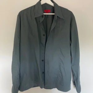 Grå Overshirt från Hugo Boss  - Stilren grå skjorta från Hugo Boss med klassisk krage och knäppning framtill. Skjortan har lång ärm och är vindtät. Perfekt för dig som gillar en clean och enkel look. Storlek L och passar dig som är runt 185cm. Saknas en knapp därav billigare pris.