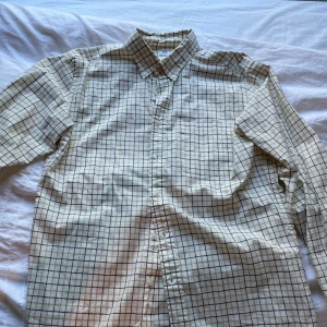 Rutig skjorta från Uniqlo, beige/brun - Klassisk rutig skjorta från Uniqlo i beige och brunt. Skjortan har button-down krage, bröstficka och knäppning framtill. Tillverkad i 100% bomull för en skön och luftig känsla. Perfekt för en clean och avslappnad stil.