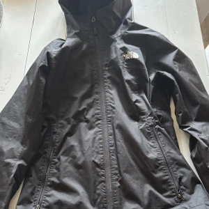 Svart vindjacka från The North Face XS - Svart vindjacka från The North Face i storlek XS. Jackan har huva, hel dragkedja framtill och två sidofickor med dragkedja. Materialet är slitstark syntet med meshfoder på insidan. Snygg logga på bröstet och ryggen. Perfekt för blåsiga dagar. Har endast använt den ett fåtal gånger💓💓