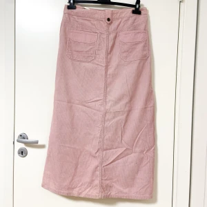 Rosa manchesterkjol från Authentic - Lång rosa manchesterkjol från Authentic Clothing Company med raka linjer, stora fickor framtill och dekorativa knappar längs sidorna. Kjolen är i 100% bomull och har coola detaljer som ger en retrovibe.