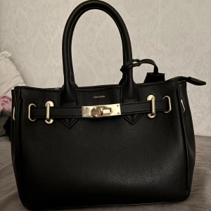 Svart handväska från Don Donna - Stilren svart handväska från Don Donna i skinnimitation med guldfärgade metalldetaljer och lås framtill. Väskan har två rejäla handtag och dragkedja upptill. Perfekt storlek för att få plats med allt du behöver och ger en lyxig känsla till din outfit.
