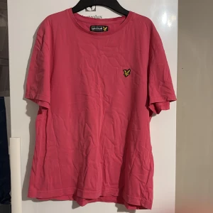 Rosa t-shirt från Lyle & Scott - Snygg rosa t-shirt från Lyle & Scott med klassisk rund hals och korta ärmar. T-shirten har en gul broderad logga på bröstet och är gjord i mjuk bomull. Perfekt för en avslappnad och stilren look.