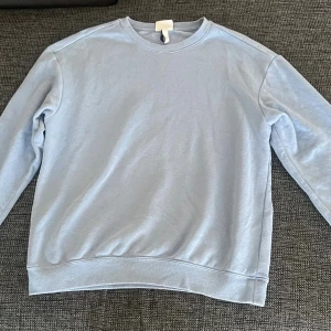 Ljusblå sweatshirt från H&M Essentials - En stilren ljusblå sweatshirt från H&M Essentials, modell No. 5. Tröjan har relaxed fit, rund hals och långa ärmar. Tillverkad i mjuk bomullsmix med ribbade muddar vid ärmslut och nederkant. Perfekt för chill dagar och enkel att matcha med jeans eller joggers.