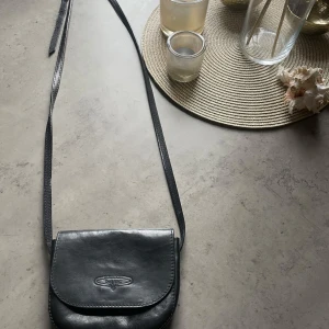 Svart axelväska från Bel Sac i skinn - Stilren svart axelväska från danska märket Belsac i äkta skinn. Väskan har ett klassiskt lock med magnetknäppning och ett huvudfack med dragkedja. Perfekt storlek för det viktigaste och enkel att bära med lång axelrem. Inga tecken på användning.