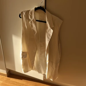 Vit lång väst från H&M - Stilren vit väst från H&M i längre modell med klassisk krage och knäppning framtill. Västen har två fickor och är tillverkad i ett lätt, slätt material som ger en clean look. Perfekt att slänga över en t-shirt eller skjorta för en snygg layering-effekt.