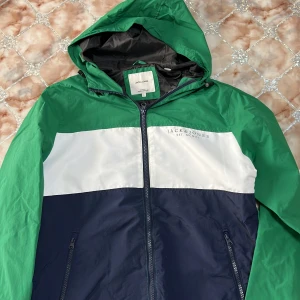 Jack & Jones vindjacka grön/blå L - Snygg vindjacka från Jack & Jones i storlek L. Jackan har färgblock i grönt, vitt och mörkblått, huva och hel dragkedja framtill. Tillverkad i lätt polyester, perfekt för blåsiga dagar. Två fickor med dragkedja och sportig look.