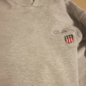 Grå zip-jacka från Gant - Säljer en stilren grå zip-jacka från Gant med broderad logga och sköld på bröstet. Jackan har hög krage och dragkedja framtill. Perfekt för dig som gillar klassisk och enkel stil. Finns i Grums tar bara cash