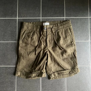 Olivgröna shorts från John Henric - Snygga olivgröna shorts från John Henric med snörning i midjan och uppvikta benslut. Tillverkade i en skön mix av bomull och linne, perfekta för varma dagar. Klassisk design med fickor fram och bak samt knapp- och dragkedjestängning.