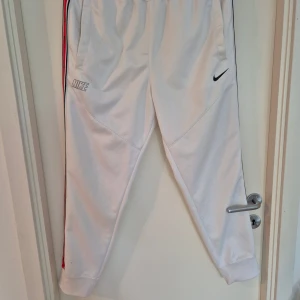 Vita mjukisbyxor från Nike med röd detalj - Snygga vita mjukisbyxor från Nike med svarta och röda ränder längs sidorna. Byxorna har elastisk midja med snörning, fickor på sidorna och Nike-logga framtill. Perfekta för chill eller träning, i mjukt bomullsmaterial.