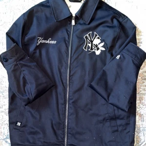 Mörkblå Yankees vindjacka med dragkedja - Snygg mörkblå Yankees vindjacka från New Era med broderad logga och text på bröstet. Jackan har klassisk krage, hel dragkedja framtill och långa ärmar. Insidan är fodrad med ljusblått, randigt tyg. Perfekt för dig som gillar sportig streetstyle.
