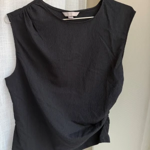 Svart ärmlös topp - En snygg svart topp med rund halsringning och lätt strukturerat tyg. Toppen är ärmlös och har en rak passform på ena sidan och med en liten innerbenssöm på sidan.