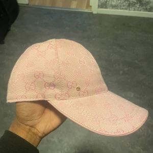 Rosa Gucci keps med monogram - Snygg rosa keps från Gucci med klassiskt GG-monogram i ljusrosa över hela kepsen. Kepsen har böjd skärm, knapp på toppen och justerbar rem bak. Tillverkad i bomull med exklusiv känsla, perfekt för dig som vill sticka ut med designerstil.