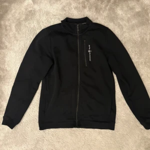 Svart ziptröja från Sail Racing - Svart långärmad ziptröja från Sail Racing med hög krage och dragkedja framtill. Tröjan har en bröstficka med dragkedja och logga tryckt i vitt på bröstet. Perfekt för lager-på-lager under kyliga dagar.